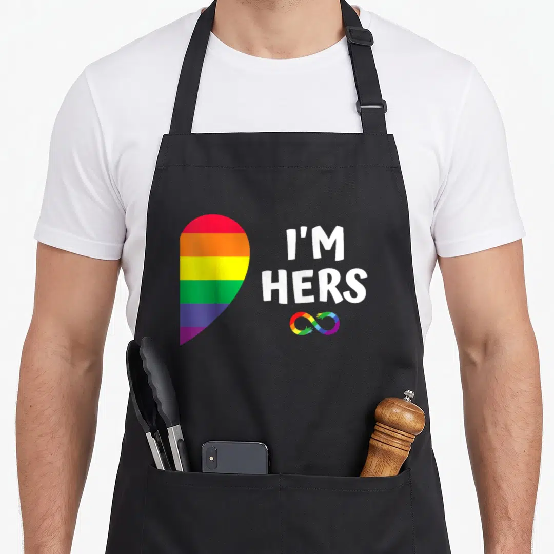 Pride she's in her apron - Rainbow heart next to white I'M HERS text and colored infinity symbol — shown on a black cotton canvas apron against a white background