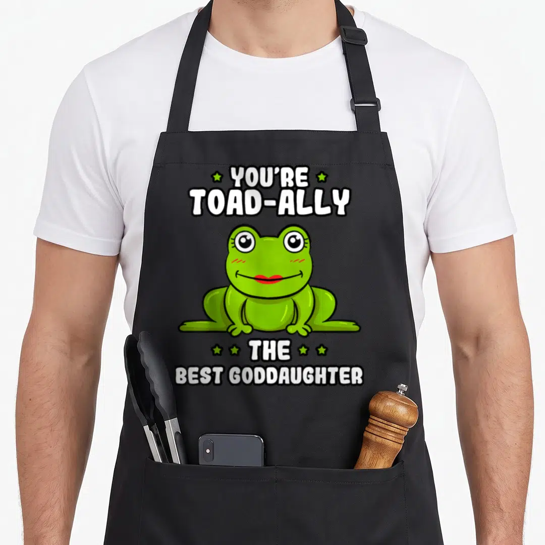 Ally frog aprons - Green cartoon frog between YOU'RE TOAD-ALLY THE BEST GODDAUGHTER text and stars — shown on a black cotton canvas apron against a white background