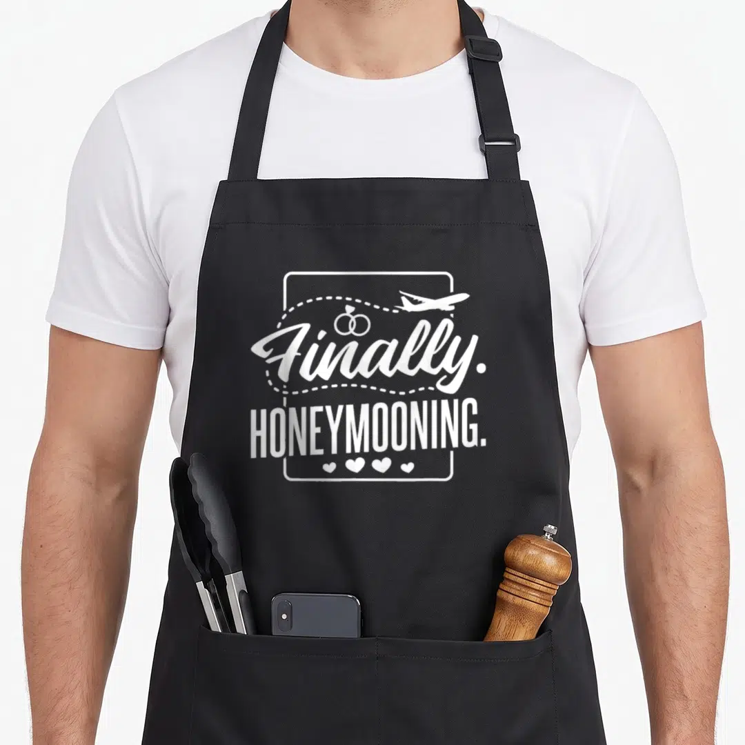 Finally newlywed aprons - White text Finally Honeymooning with plane rings and four hearts in frame — shown on a black cotton canvas apron against a white background