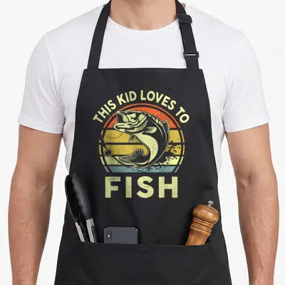 Youth fish apron - Retro sun graphic with jumping bass and THIS KID LOVES TO FISH text in yellow orange red blue — shown on a black cotton canvas apron against a white background