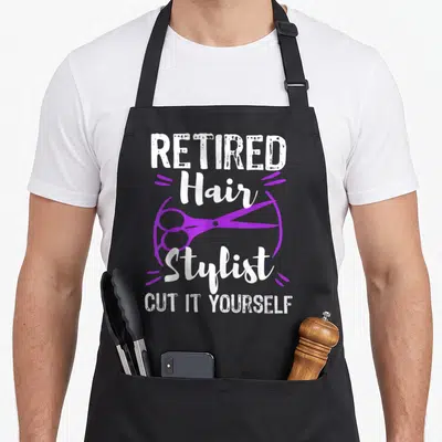 Stylist funny retirement aprons - White text reading Retired Hair Stylist Cut It Yourself with purple scissors — shown on a black cotton canvas apron against a white background