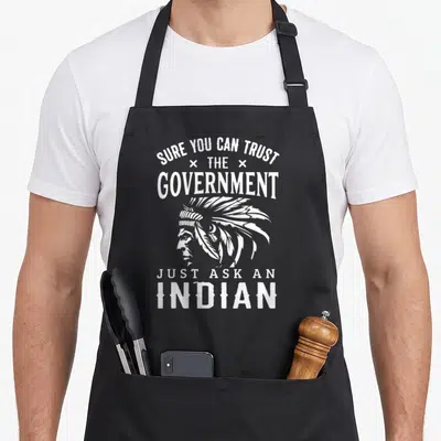Trust native american apron - White line-art of a man in profile wearing a headdress, surrounded by text reading Sure — shown on a black cotton canvas apron against a white background