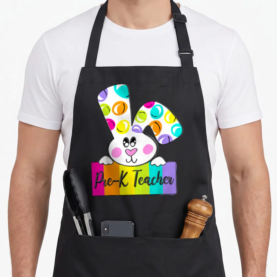 Pre easter bunny aprons - Cartoon bunny with patterned ears peeking over rainbow banner reading Pre-K Teacher — shown on a black cotton canvas apron against a white background