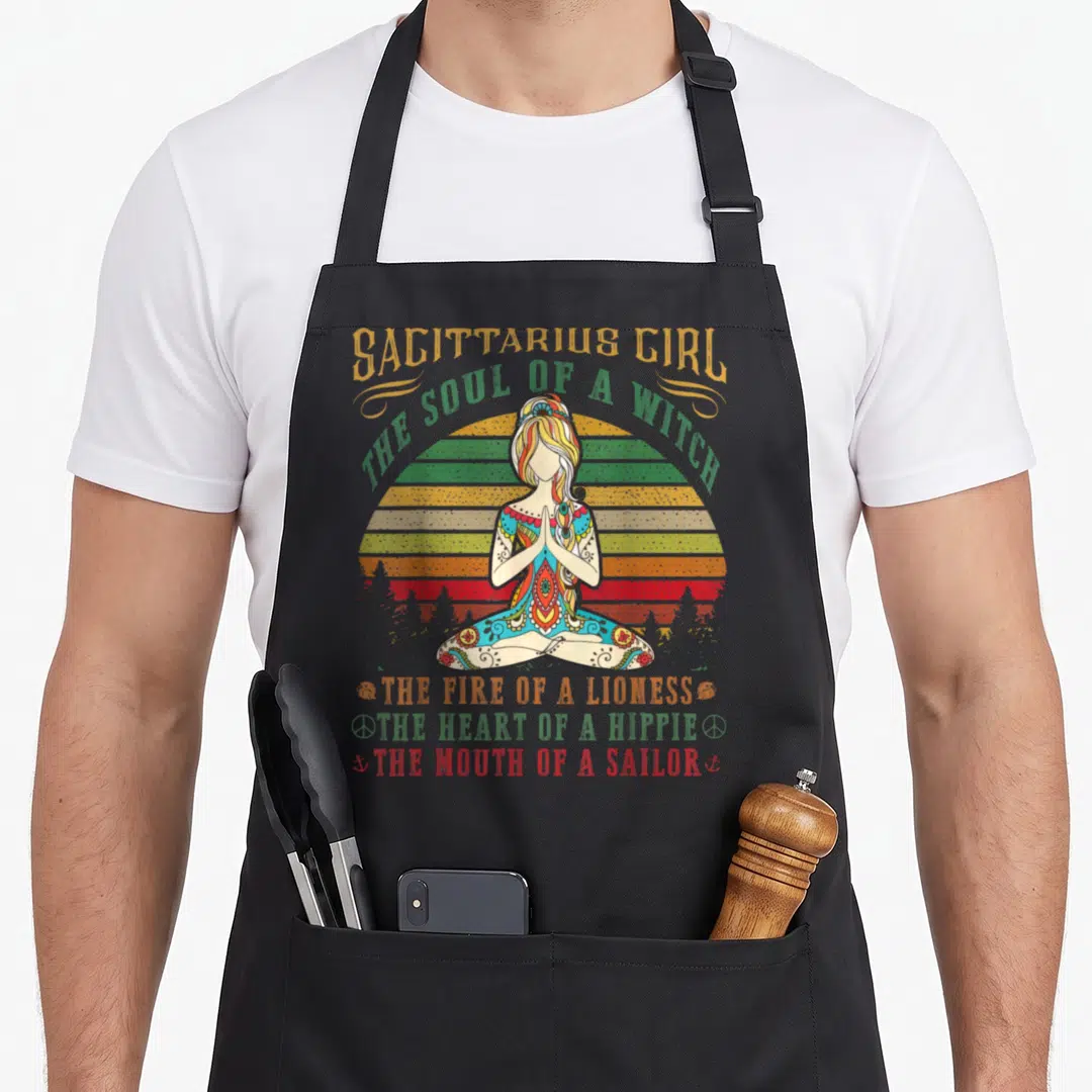 Sagittarius birthday apron - Woman in lotus pose with striped sun and text about witch lioness hippie sailor traits — shown on a black cotton canvas apron against a white background