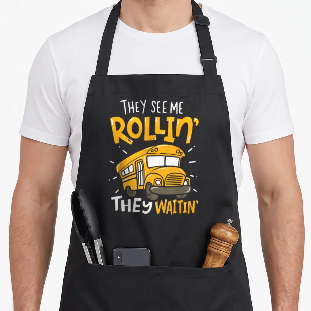 School see me aprons - Yellow cartoon school bus with THEY SEE ME ROLLIN THEY WAITIN text and white motion lines — shown on a black cotton canvas apron against a white background