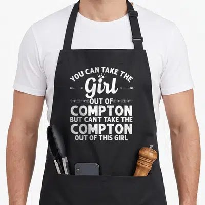 Root compton apron - Distressed white text You Can Take The Girl Out Of Compton with crown and floral dividers on black — shown on a black cotton canvas apron against a white background