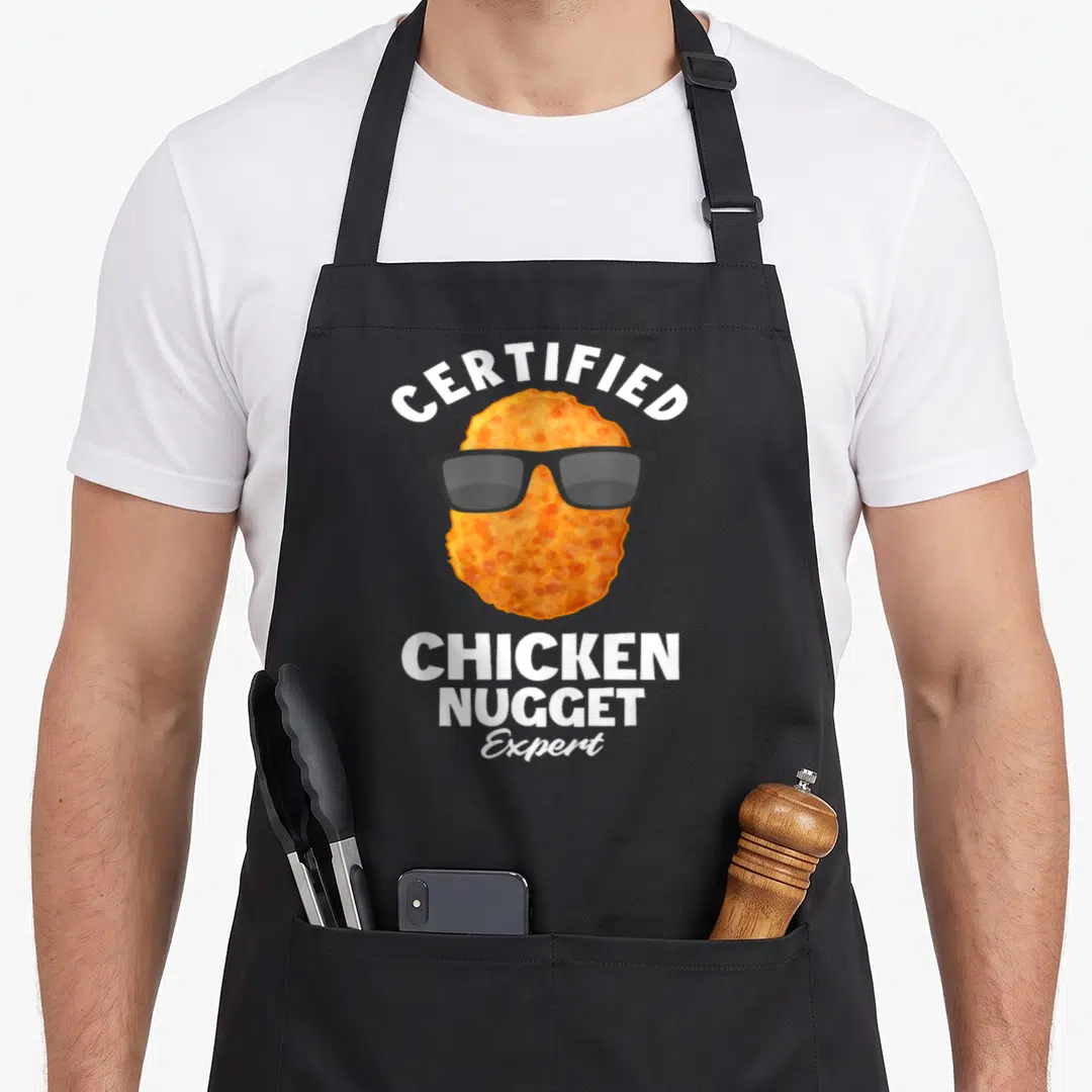 Certified chicken print apron - Cartoon orange-brown nugget wearing black sunglasses with certified chicken nugget — shown on a black cotton canvas apron against a white background
