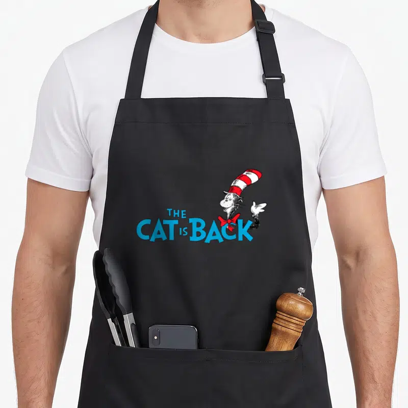 General cat apron for women - Cartoon cat with red white striped hat next to blue stacked text reading THE CAT IS BACK — shown on a black cotton canvas apron against a white background