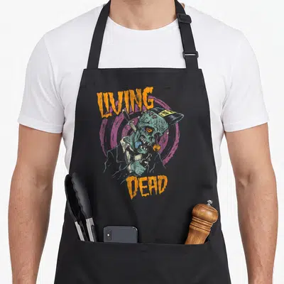 Psychobilly punk rock apron - Teal zombie on purple circle with orange LIVING DEAD text in distressed font — shown on a black cotton canvas apron against a white background