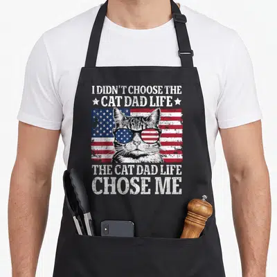 Flag cat father day apron - Distressed USA flag background with cat in sunglasses and white text reading I didn't — shown on a black cotton canvas apron against a white background
