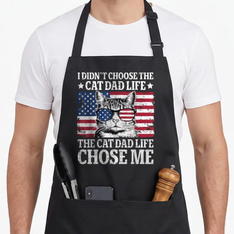 Flag cat father day apron - Distressed USA flag background with cat in sunglasses and white text reading I didn't — shown on a black cotton canvas apron against a white background