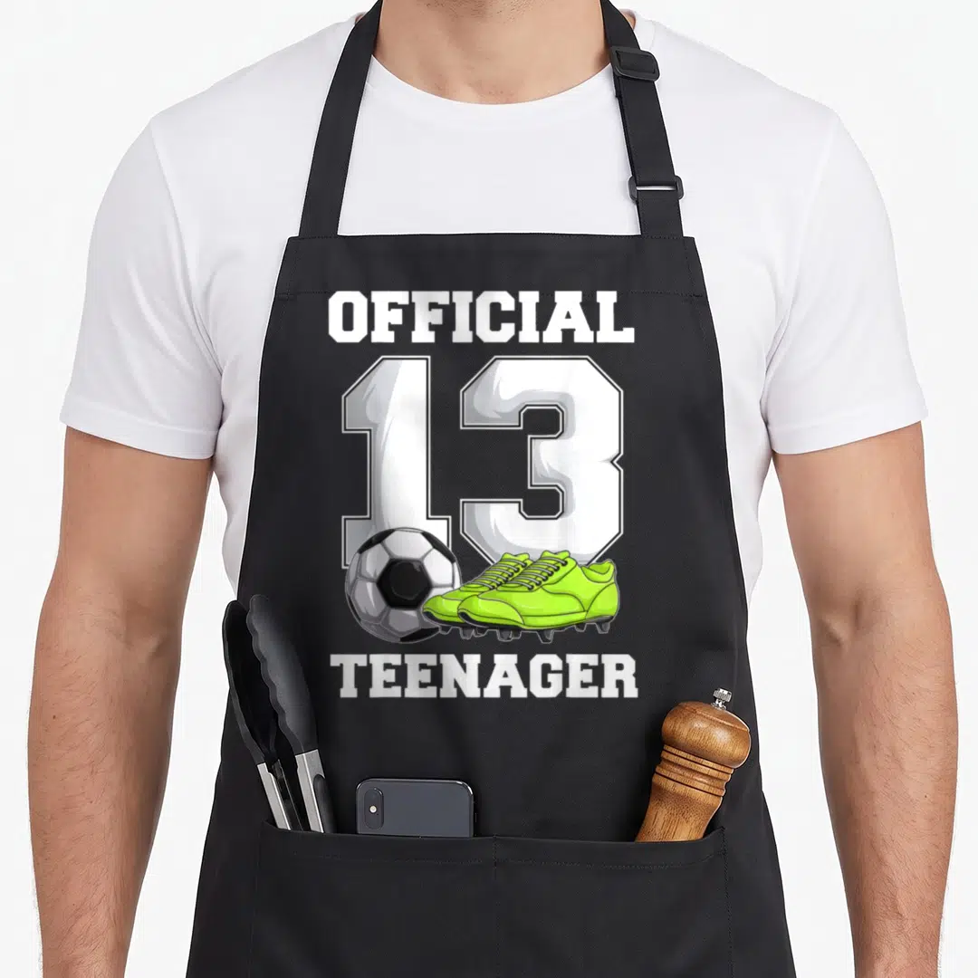 Cool soccer birthday apron - Official 13 Teenager text with soccer ball and lime green cleats in cartoon style — shown on a black cotton canvas apron against a white background
