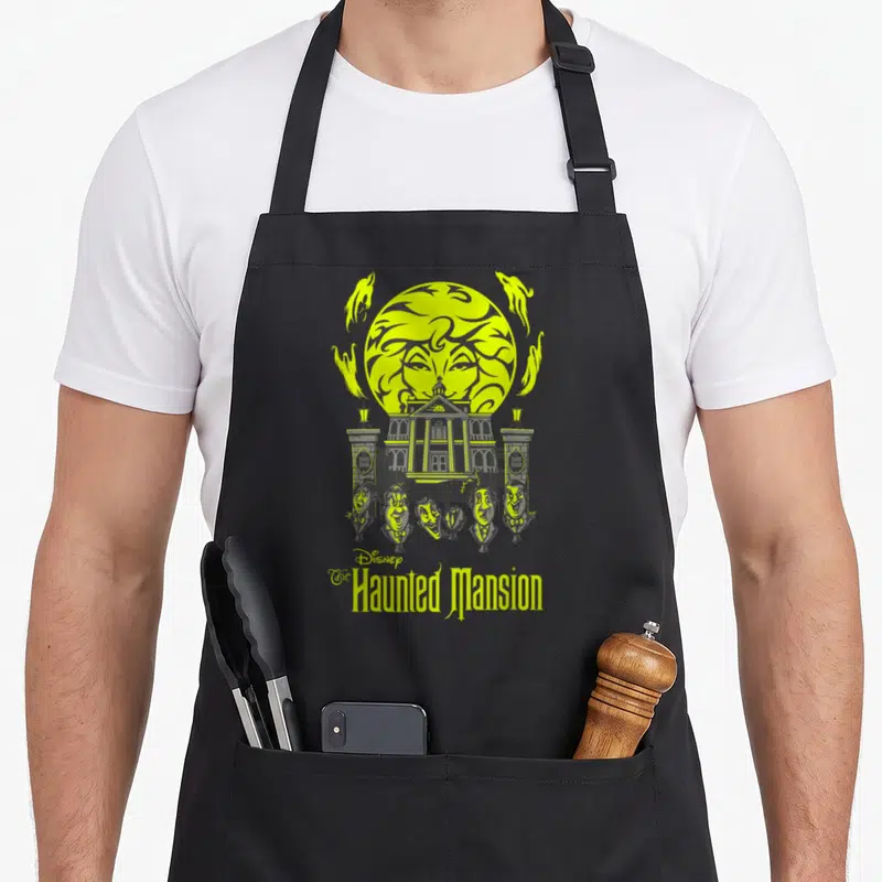 Madame haunted mansion apron - Lime green moon with face behind grey mansion and five singing busts — shown on a black cotton canvas apron against a white background