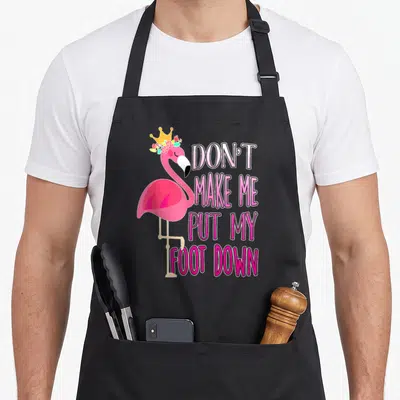 Funny awareness flamingo apron - Pink flamingo with crown and flowers next to stacked text saying DON'T MAKE ME PUT MY — shown on a black cotton canvas apron against a white background