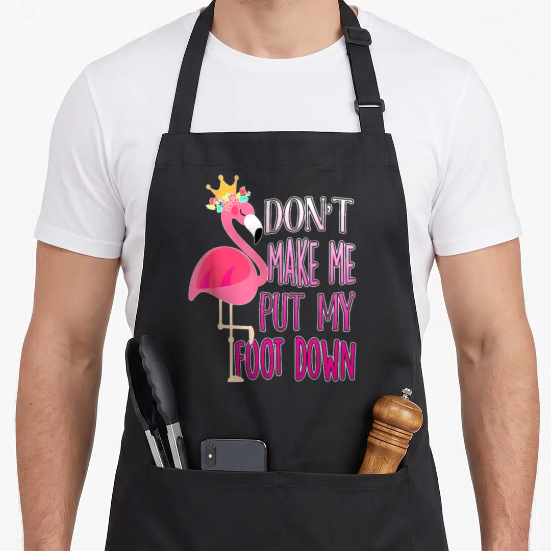 Funny awareness flamingo apron - Pink flamingo with crown and flowers next to stacked text saying DON'T MAKE ME PUT MY — shown on a black cotton canvas apron against a white background
