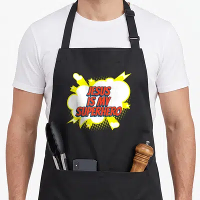 Superhero church christian apron - JESUS IS MY SUPERHERO text in red blue inside white speech bubble with yellow stars — shown on a black cotton canvas apron against a white background