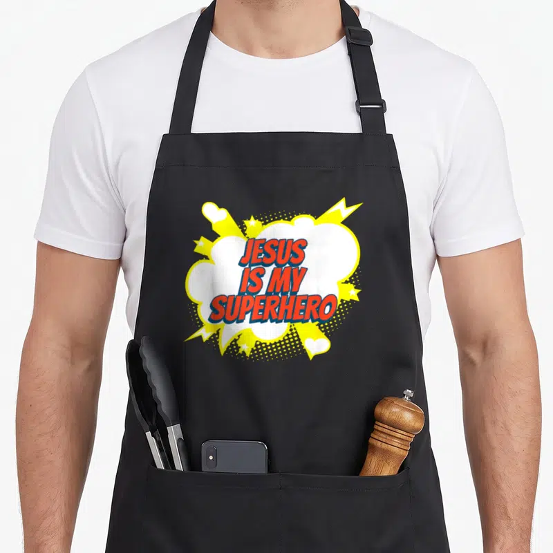 Superhero church christian apron - JESUS IS MY SUPERHERO text in red blue inside white speech bubble with yellow stars — shown on a black cotton canvas apron against a white background