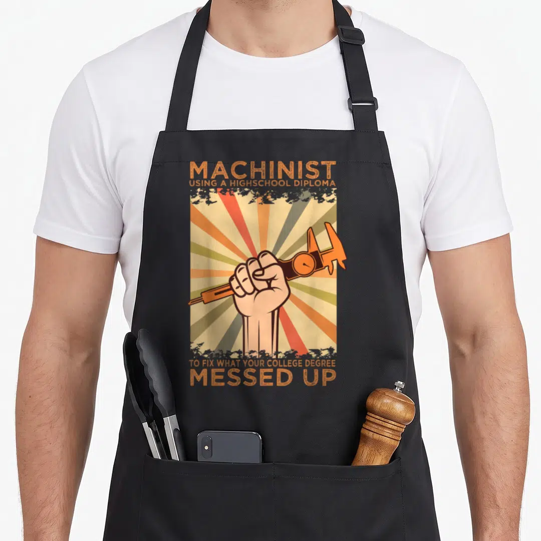 Diploma machinist apron - Retro fist holding caliper with orange sunburst and sarcastic machinist text — shown on a black cotton canvas apron against a white background