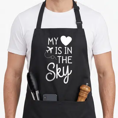 Flight sky apron - White text MY IS IN THE SKY with heart and airplane icon, vertical stack layout — shown on a black cotton canvas apron against a white background