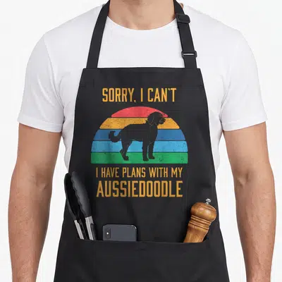 Aussiedoodle dog apron - Retro rainbow sunset with grey dog silhouette and orange text reading sorry i can't i have — shown on a black cotton canvas apron against a white background