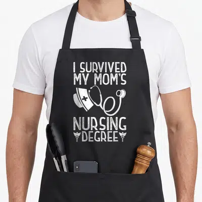 Survived nursing aprons - White distressed text I Survived My Moms Nursing Degree with nurse cap stethoscope — shown on a black cotton canvas apron against a white background