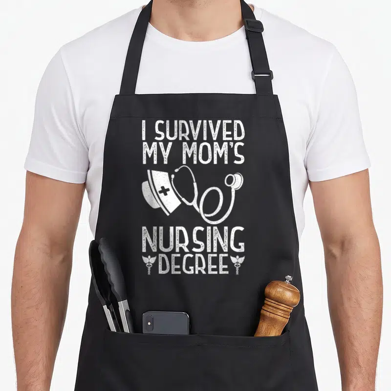 Survived nursing aprons - White distressed text I Survived My Moms Nursing Degree with nurse cap stethoscope — shown on a black cotton canvas apron against a white background