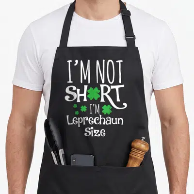 Leprechan st patrick day aprons - I'M NOT SHORT I'M Leprechaun Size text with green shamrocks replacing letters — shown on a black cotton canvas apron against a white background