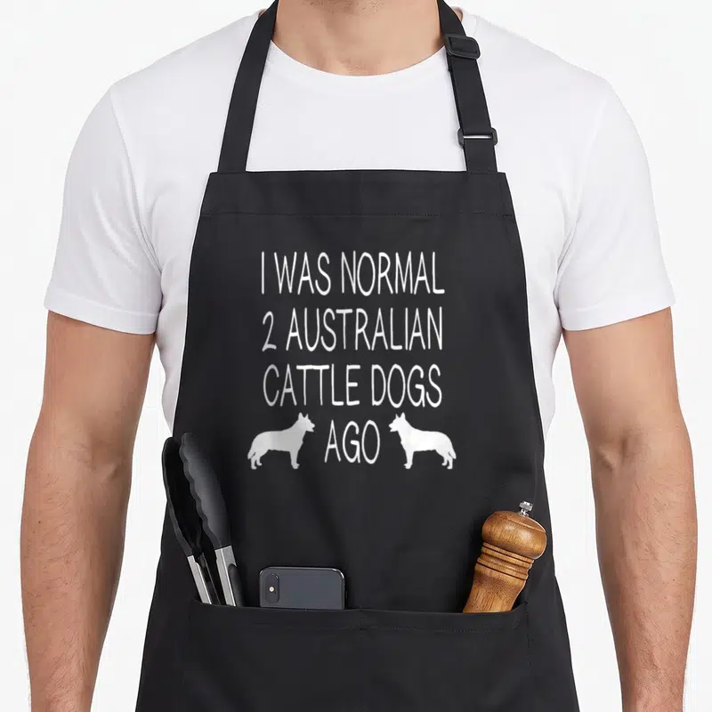 Funny australian dog kitchen apron - I WAS NORMAL 2 AUSTRALIAN CATTLE DOGS AGO text with two white dog silhouettes — shown on a black cotton canvas apron against a white background