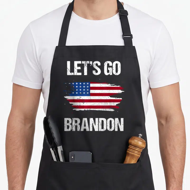 Chant novelty apron - Distressed USA flag brushstroke between LET'S GO and BRANDON text in white, red, and blue — shown on a black cotton canvas apron against a white background