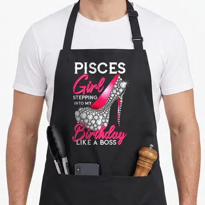 Pisce bbq boss apron - Pink rhinestone high heel shoe with Pisces birthday text — shown on a black cotton canvas apron against a white background