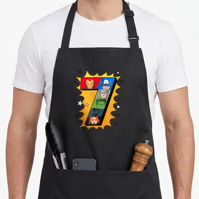 Comic holiday apron - Number seven filled with six superhero faces on yellow comic burst background — shown on a black cotton canvas apron against a white background