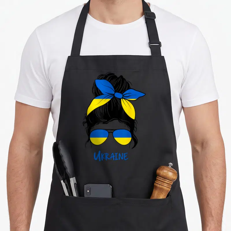 Funny ukraine ukrainian aprons - Woman head with blue yellow sunglasses headband and Ukraine text below — shown on a black cotton canvas apron against a white background