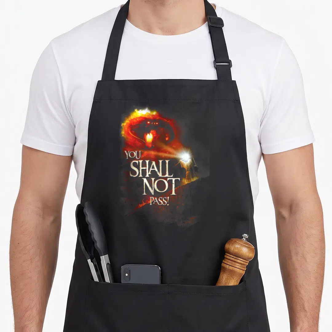 Balrog ring apron - Fiery demon head with You Shall Not Pass text and wizard casting light beam — shown on a black cotton canvas apron against a white background