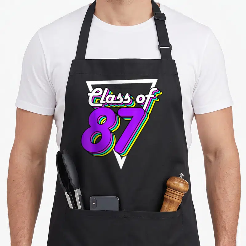 School class reunion apron - Class of 87 text with colorful 3D shadows inside a white triangle outline — shown on a black cotton canvas apron against a white background