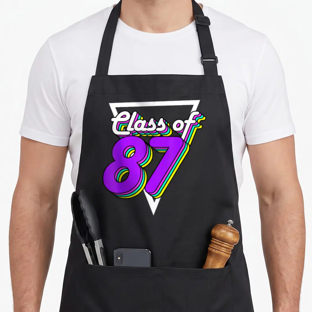 School class reunion apron - Class of 87 text with colorful 3D shadows inside a white triangle outline — shown on a black cotton canvas apron against a white background