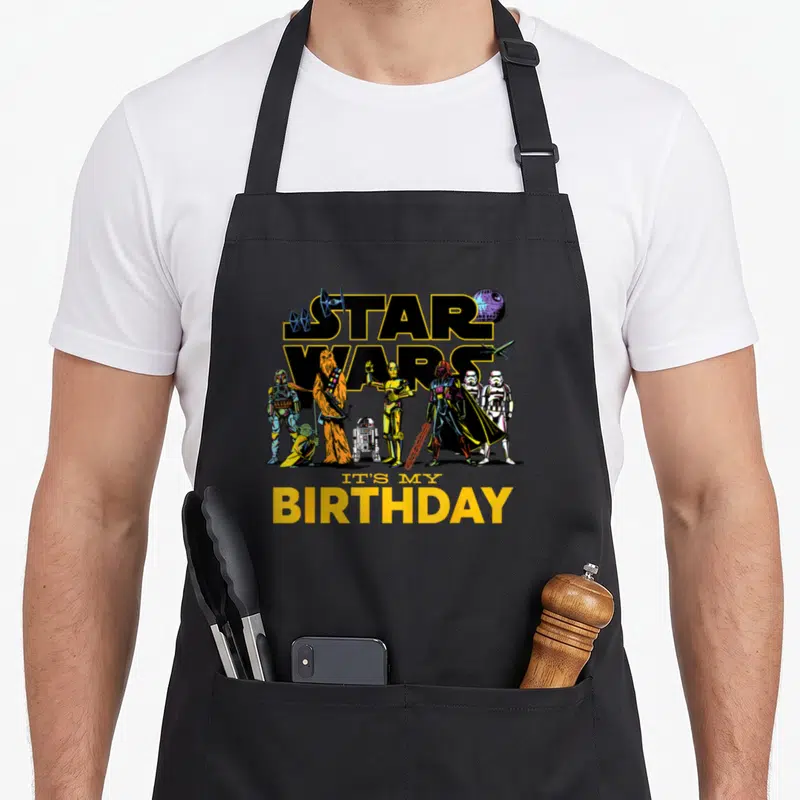 Character birthday apron - Star Wars logo with characters and It's My Birthday text in yellow black blue red — shown on a black cotton canvas apron against a white background