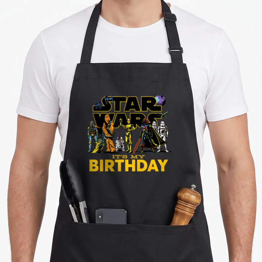 Character birthday apron - Star Wars logo with characters and It's My Birthday text in yellow black blue red — shown on a black cotton canvas apron against a white background
