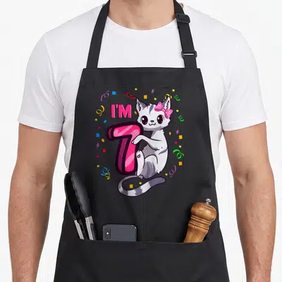 7th kitty cat apron - Cartoon cat leaning on pink number 7 with confetti — shown on a black cotton canvas apron against a white background