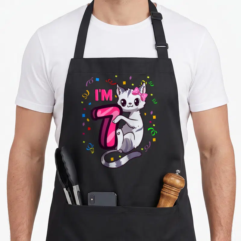 7th kitty cat apron - Cartoon cat leaning on pink number 7 with confetti — shown on a black cotton canvas apron against a white background