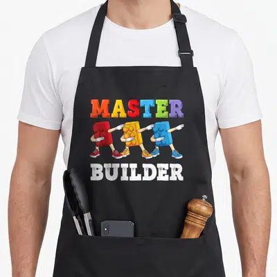 Builder building block & apron - Colorful MASTER BUILDER text with three walking toy bricks in red yellow blue white — shown on a black cotton canvas apron against a white background