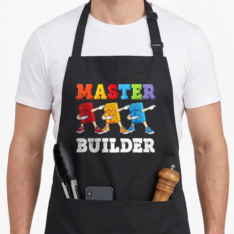 Builder building block & apron - Colorful MASTER BUILDER text with three walking toy bricks in red yellow blue white — shown on a black cotton canvas apron against a white background