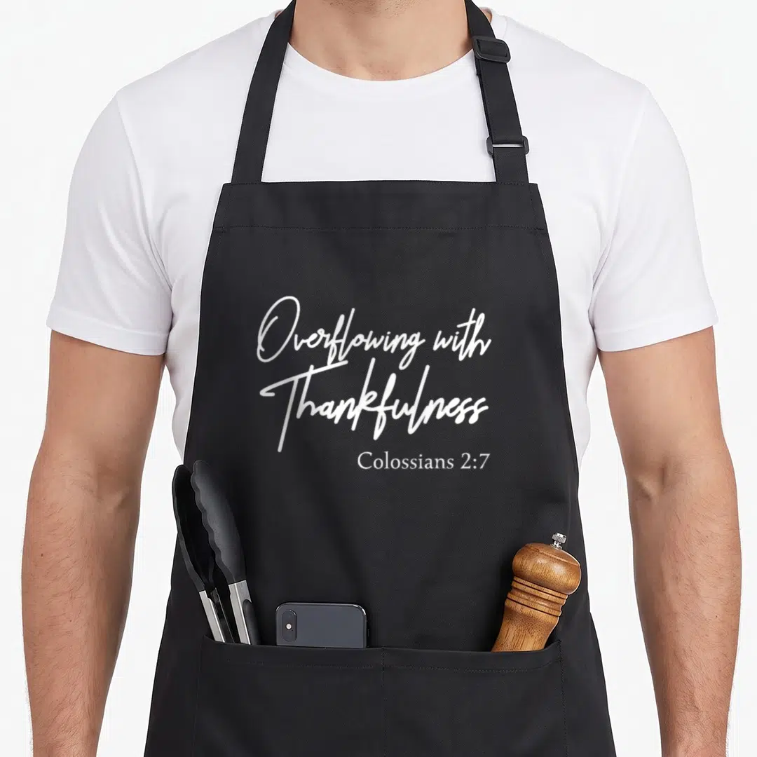 Colossian thanksgiving apron - White script text Overflowing with Thankfulness and Colossians 2:7 on grey background — shown on a black cotton canvas apron against a white background