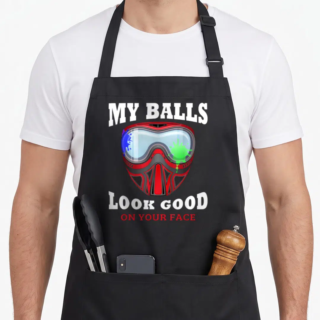 Ball face apron - Red paintball mask with blue green splatters and white text MY BALLS LOOK GOOD ON YOUR FACE — shown on a black cotton canvas apron against a white background
