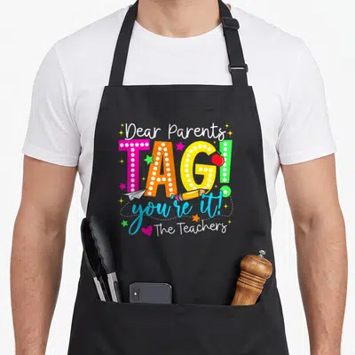 Dear tag apron - Colorful text reading Dear Parents TAG you're it The Teachers with apple and pencil icons — shown on a black cotton canvas apron against a white background