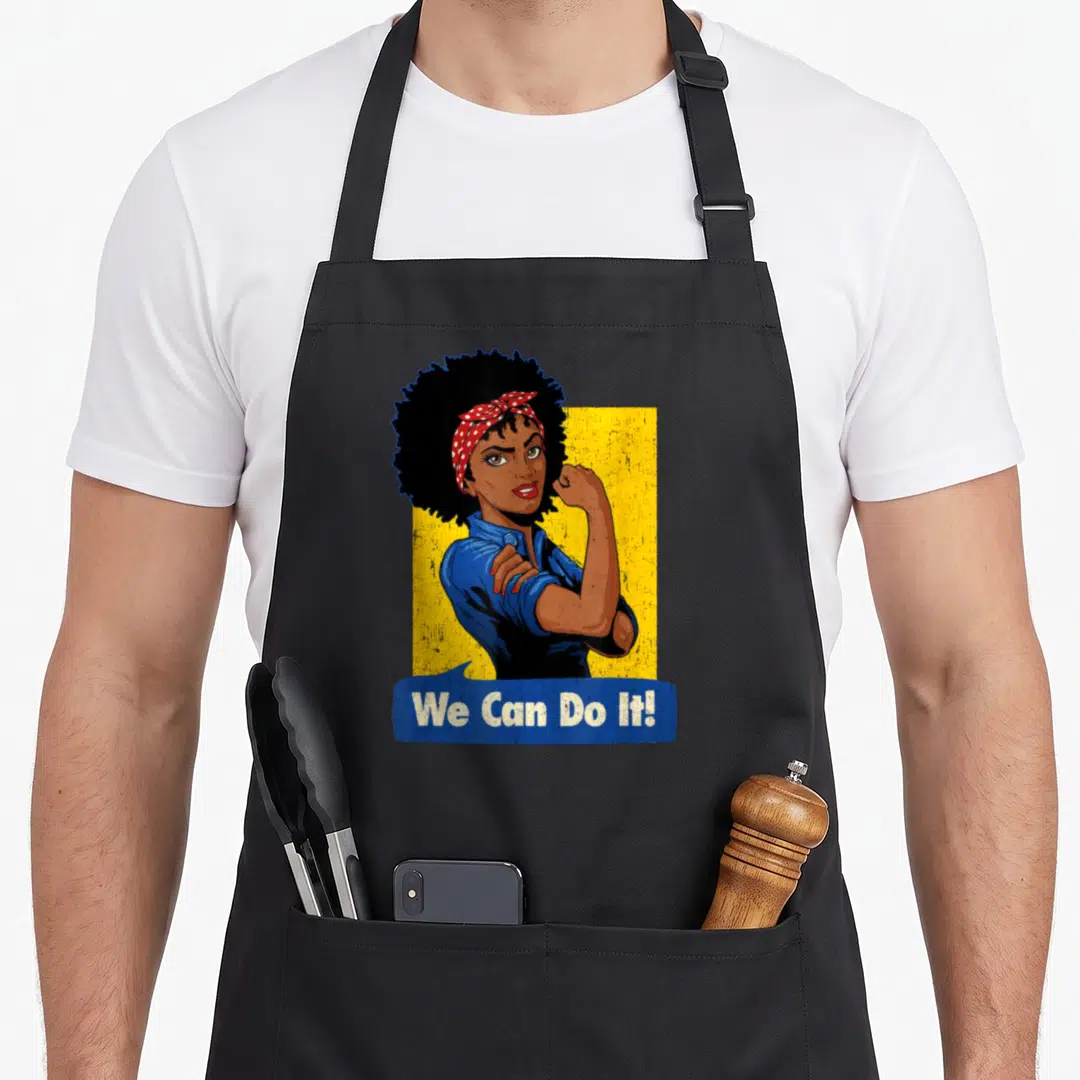 Strong rosie the riveter apron - Woman with afro and red bandana flexing arm in front of yellow background with blue — shown on a black cotton canvas apron against a white background