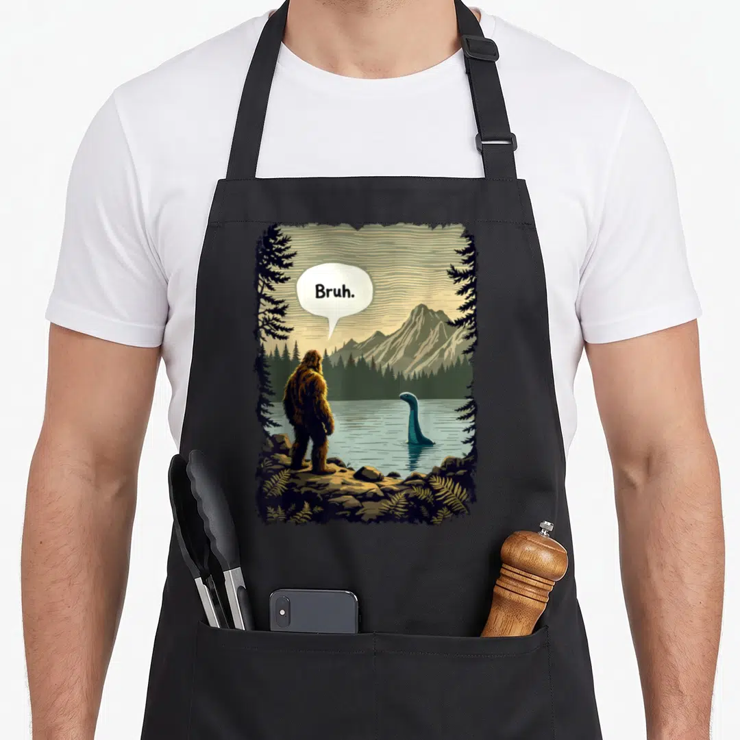 Bruh bigfoot apron - Cartoon Bigfoot standing on rocks saying Bruh to a blue-grey lake monster near mountains — shown on a black cotton canvas apron against a white background