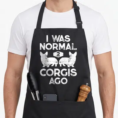 Normal corgis dog apron - White distressed text I WAS NORMAL 2 CORGIS AGO with two facing corgi silhouettes and number — shown on a black cotton canvas apron against a white background