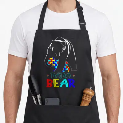 Awareness bear apron - White bear outline holding colorful puzzle cub with PAPA BEAR text — shown on a black cotton canvas apron against a white background