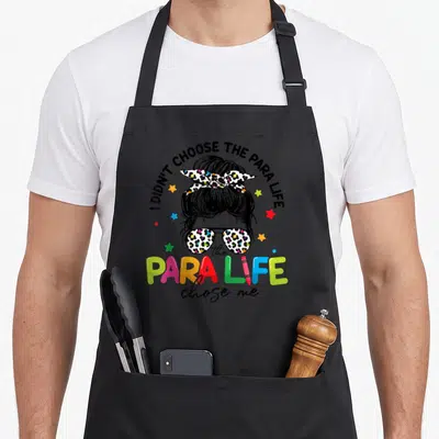 Para teacher apron - Cartoon head with bun and sunglasses surrounded by I didn't choose the para life text and colorful — shown on a black cotton canvas apron against a white background