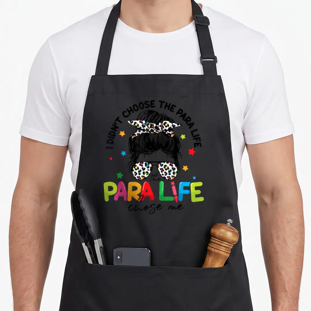 Para teacher apron - Cartoon head with bun and sunglasses surrounded by I didn't choose the para life text and colorful — shown on a black cotton canvas apron against a white background